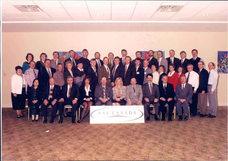 A group photo of approximately 40 people, some sitting and some standing, in formal attire in front of a NAV CANADA sign indoors.